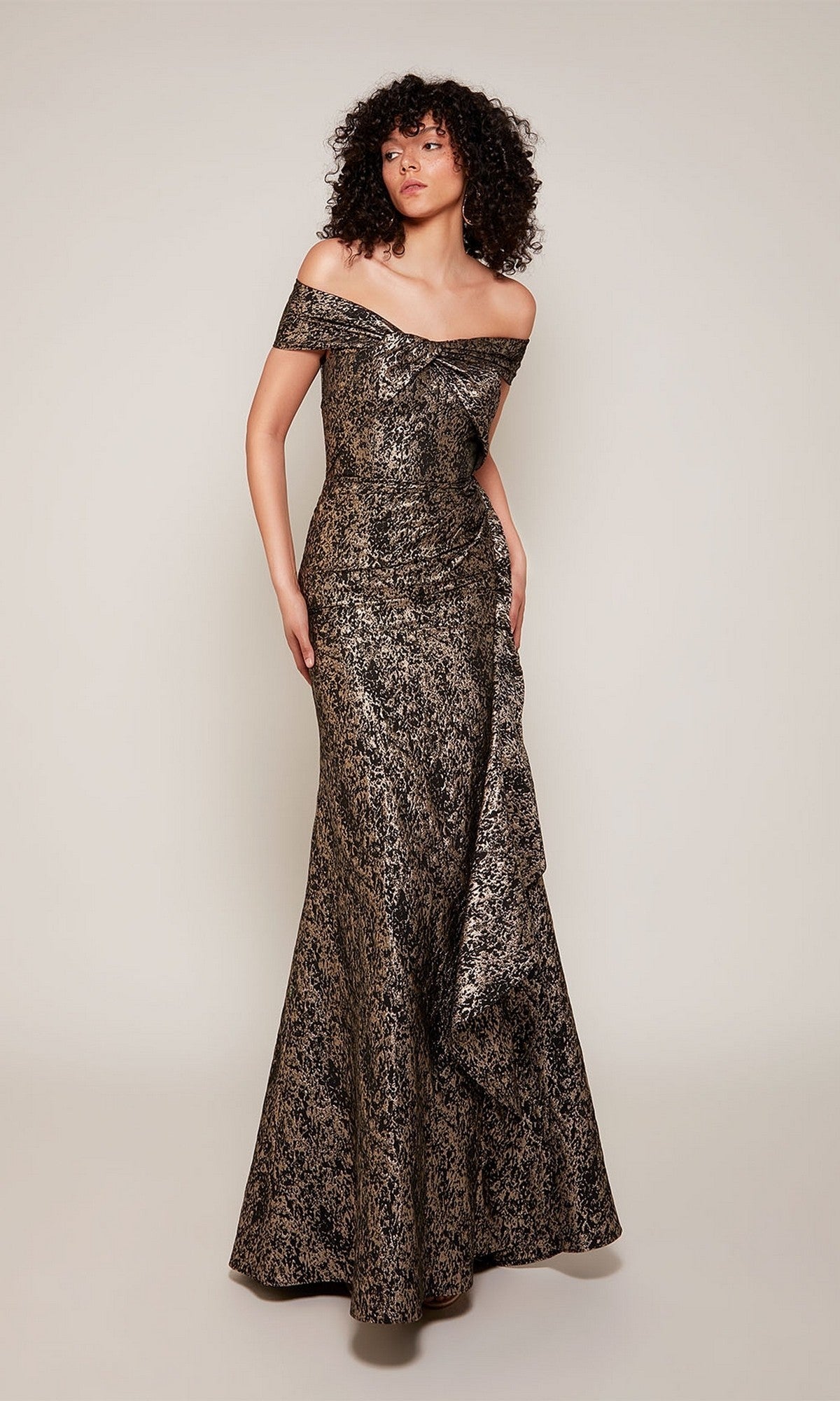 A person stands against a plain background wearing the Alyce 27667, a long formal off-the-shoulder metallic jacquard dress with a slight train—perfect for formal events or as a mother-of-the-bride gown.